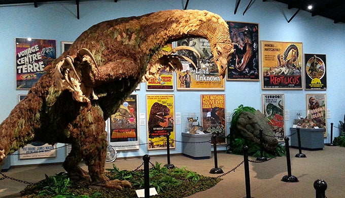UT-Blanding-Dinosaur-Museum-2_Gloria_680 A life-size feathered dinosaur model in front of the international display of dinosaur movie posters at The Dinosaur Museum.